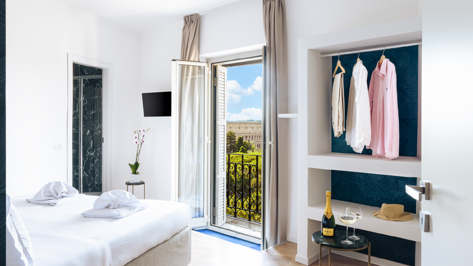 Suite Deluxe con letto matrimoniale, bagno con rivestimento scuro, balconcino aperto e Colosseo visibile sullo sfondo.