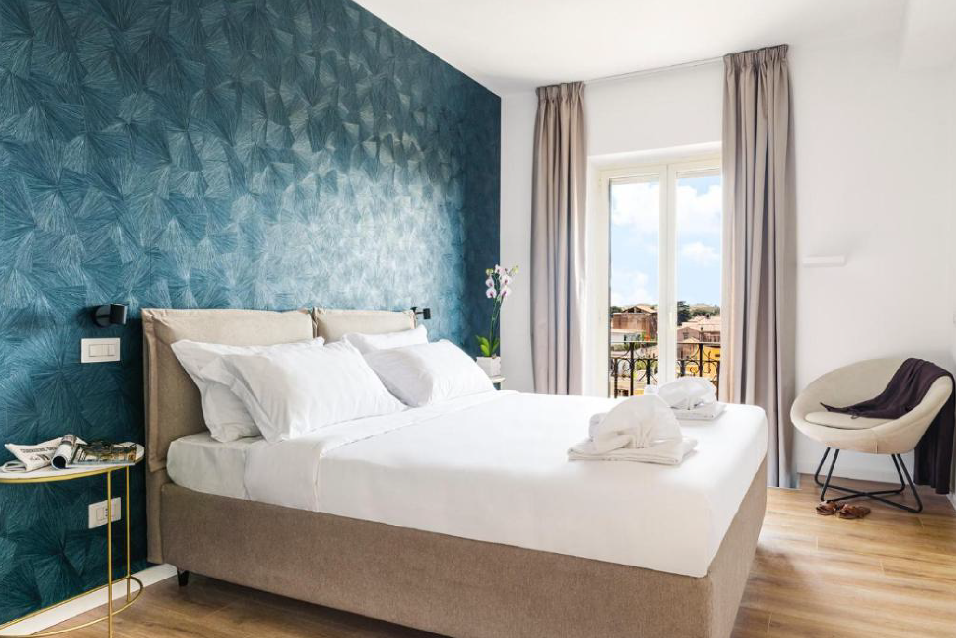 Inquadratura della camera con letto matrimoniale, parete blu, portafinestra con tende e balconcino, poltrona chiara in angolo.