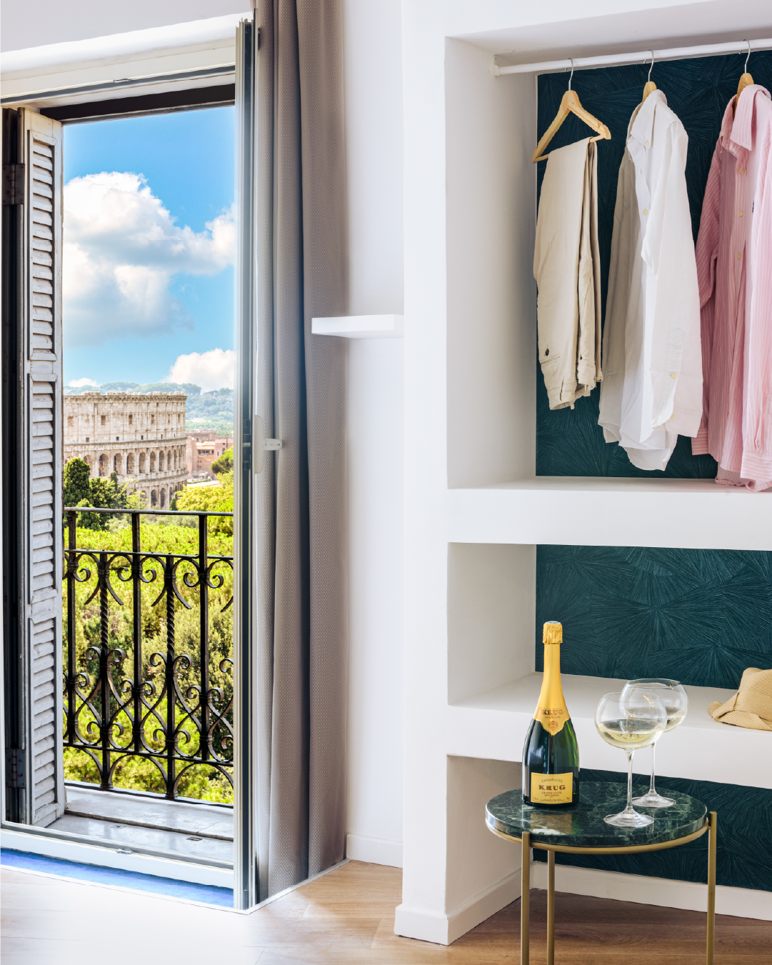 Dettaglio nicchia guardaroba con abiti appesi e tavolino con champagne e calici vicino alla portafinestra con vista Colosseo.