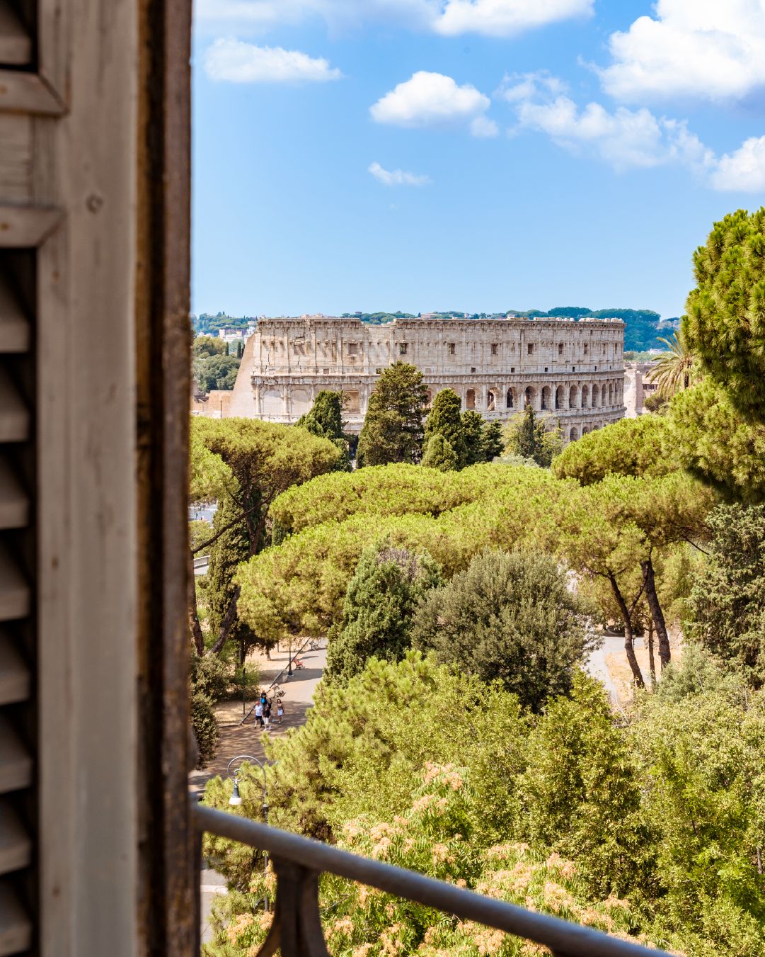 Vista sul Colosseo sopra il verde del parco, inquadrata da una finestra con persiane e ringhiera del balcone.