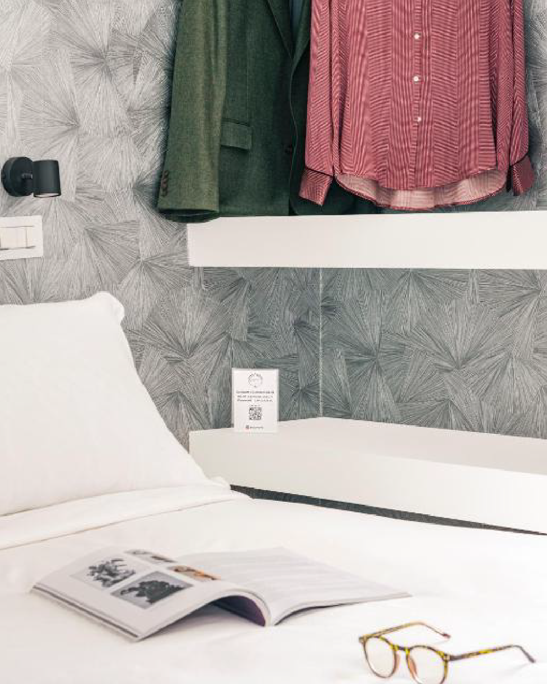 Dettaglio camera con letto matrimoniale, mensole e guardaroba a vista, libro e occhiali sul letto, parete con motivo decorativo.